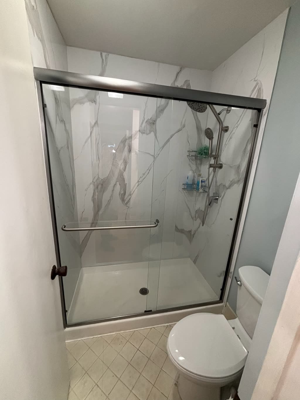 A finished walk-in shower where a bathtub used to be, with white marble-look wall panels, a low white shower base, a stainless grab bar and a sliding glass door in a bronze frame.